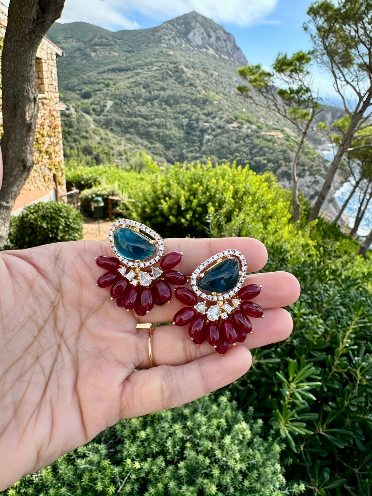 Red and blue earrings held in a hand with a scenic background